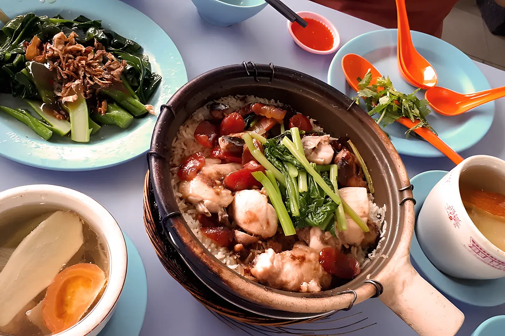 Claypot chicken rice with greens, sausage, and broth on a table. Bright orange spoons, small chili sauce bowl, and leafy greens garnish complete the setup.