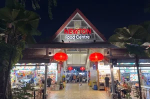 A bustling nighttime view of the Newton Food Centre entrance in Singapore, featuring its iconic triangular roof, illuminated red lanterns, and lively hawker stalls.