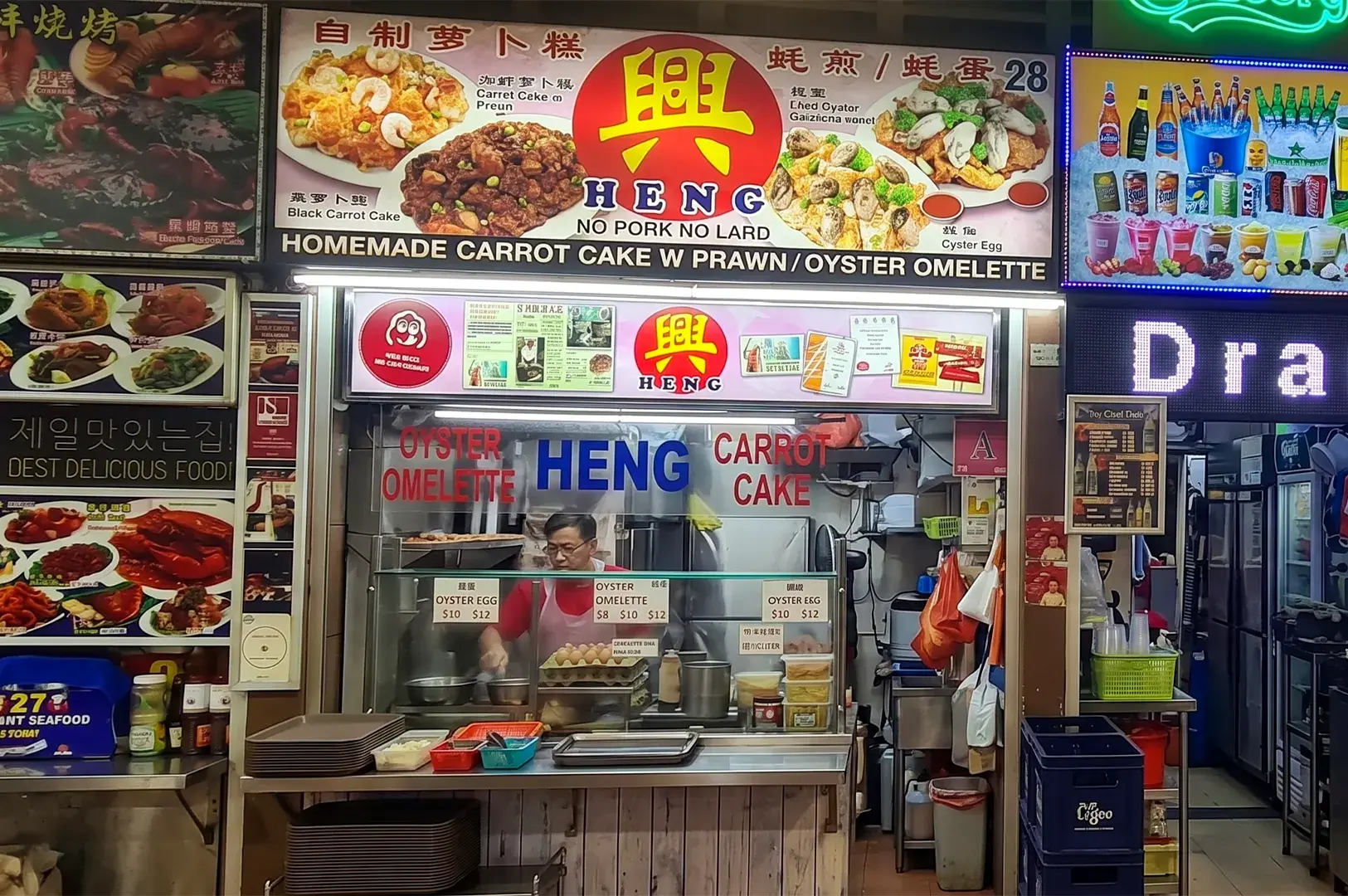 A vendor prepares food at the Heng hawker stall, which specializes in homemade carrot cake and oyster omelettes, as indicated by the large colorful signage above.
