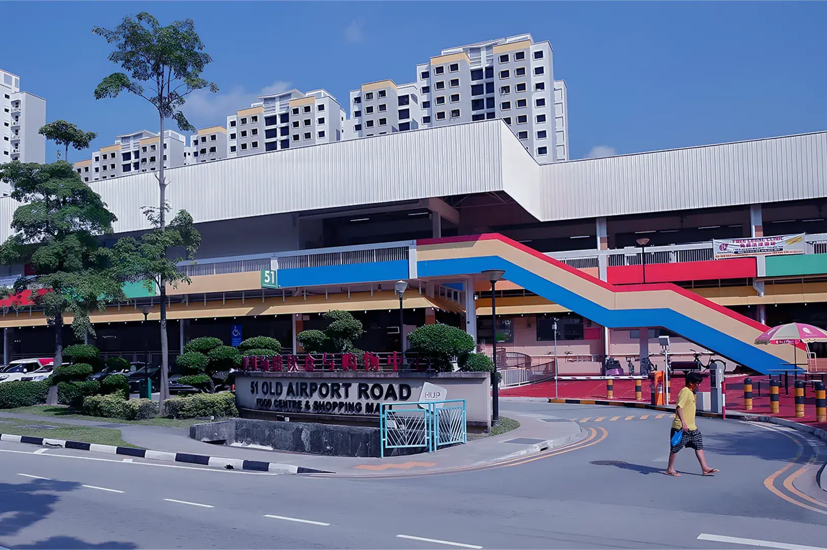 A colorful, urban scene of Old Airport Road Food Centre in Singapore, featuring a vibrant red-and-blue staircase, modern apartments, and a person walking.