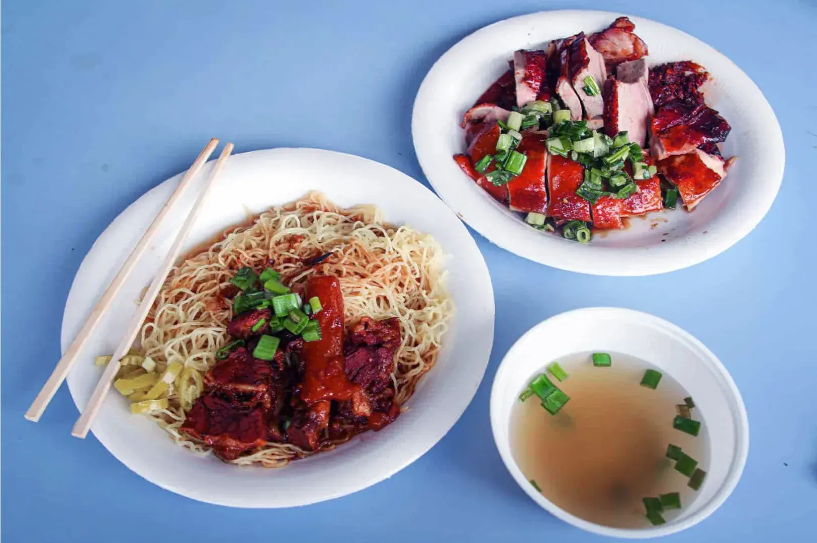 A vibrant meal with three plates on a blue table: noodles with roast duck and green onions; sliced roast meat topped with green onions; and a clear broth soup with scallions.