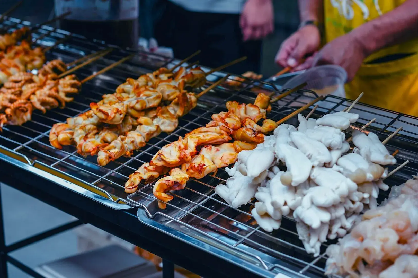 Skewered street food, including grilled meat and white seafood, on a sizzling metal grill. A vendor in a yellow apron holds tongs.