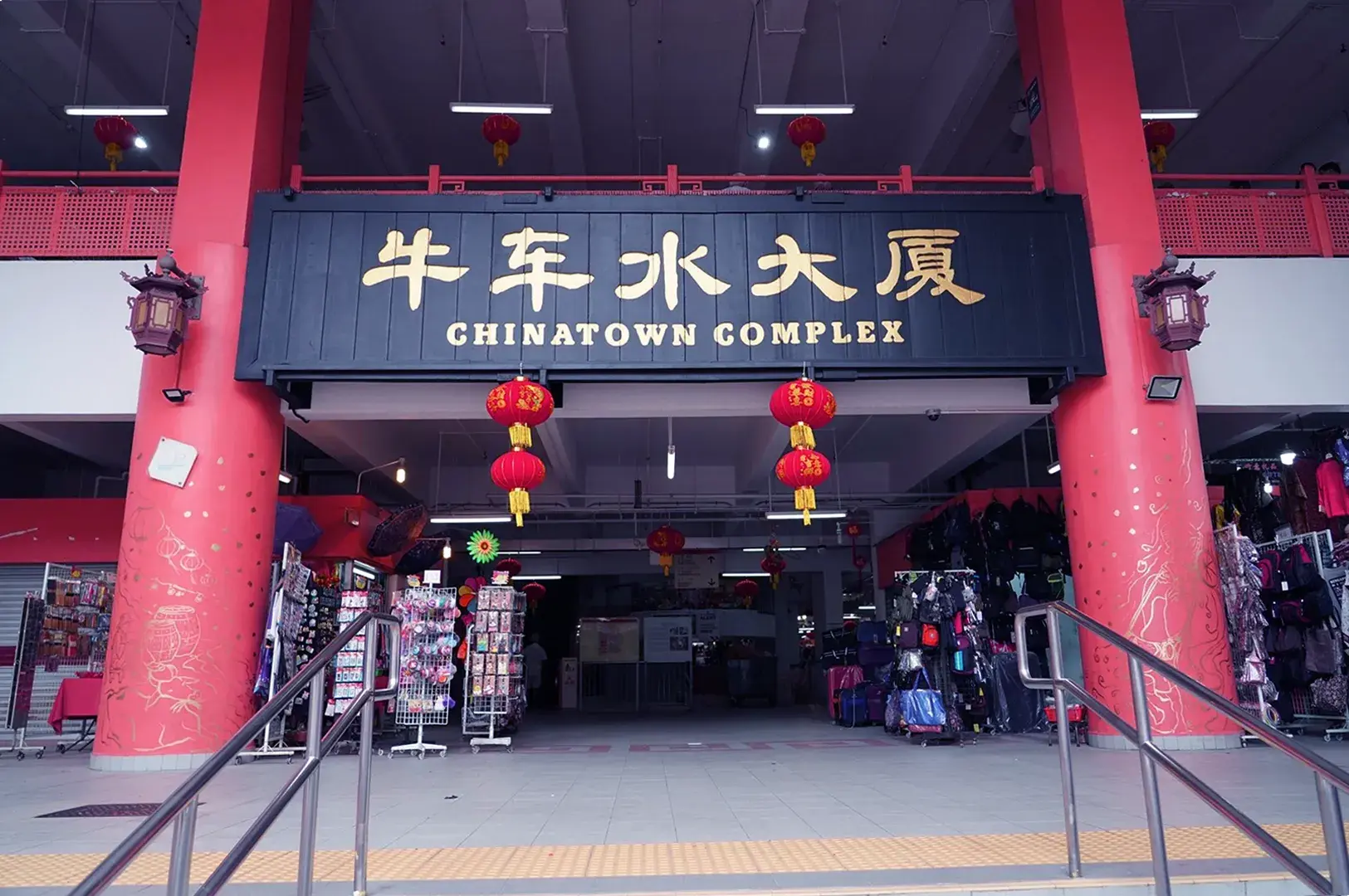 Entrance to Chinatown Complex adorned with red lanterns. Flanked by shops with vibrant merchandise. Sign in Chinese and English above. Festive ambiance.