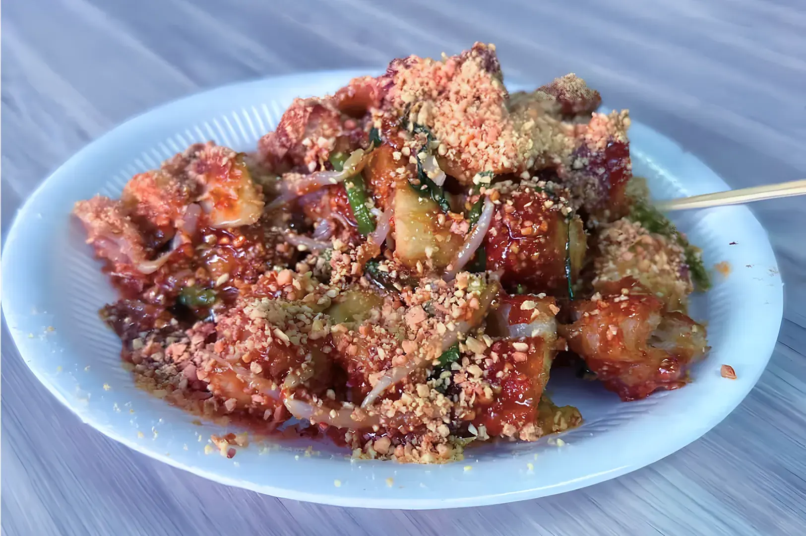 A vibrant plate of rojak on a white dish features assorted fruits and vegetables coated in a rich, spicy sauce, sprinkled with crushed peanuts.