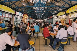 Busy indoor food market with people seated at yellow tables, surrounded by vibrant food stalls. The atmosphere is lively and bustling.