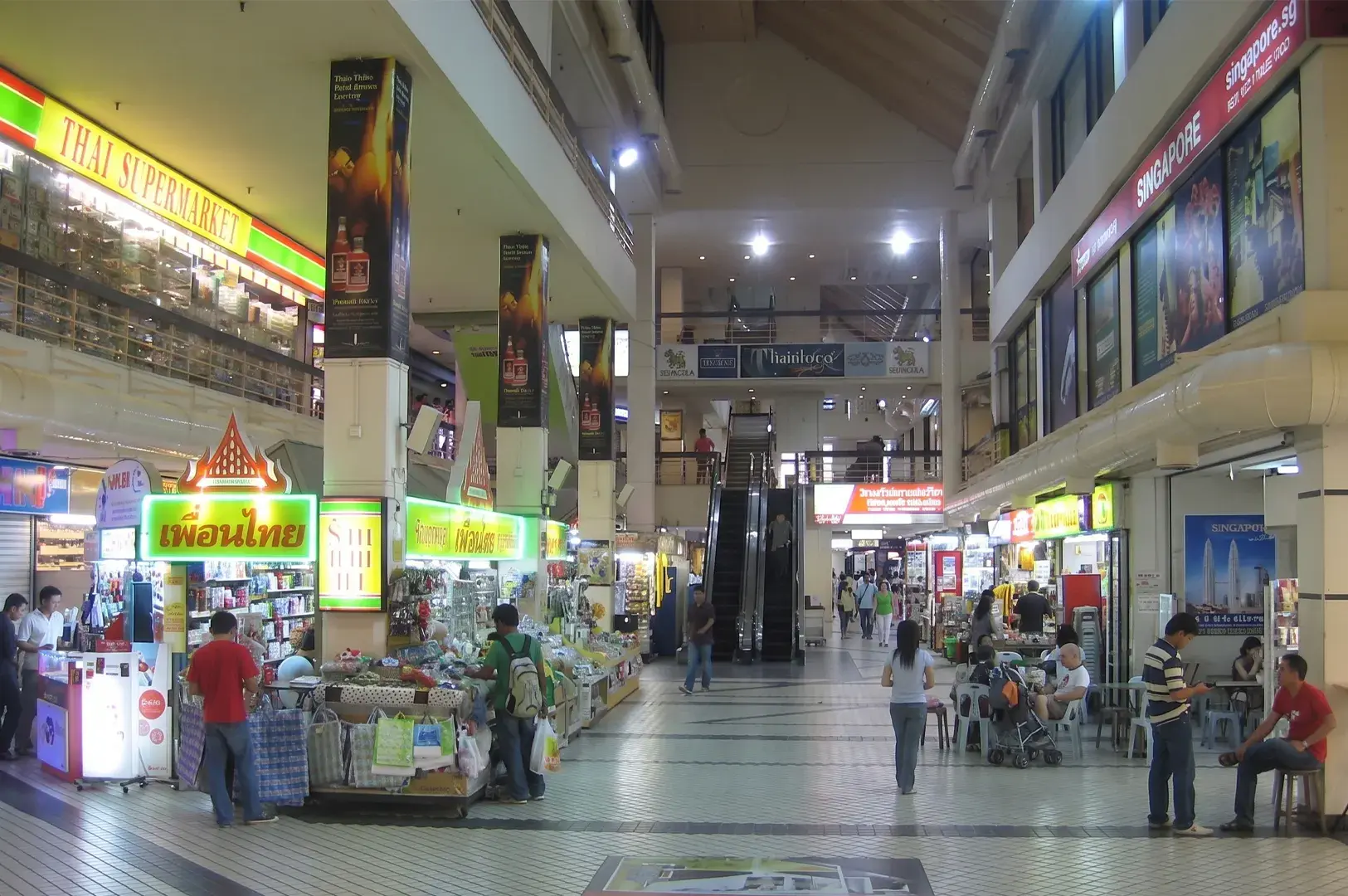 Interior of a multi-level shopping mall featuring a Thai supermarket, various small retail stalls, and an escalator in the center.