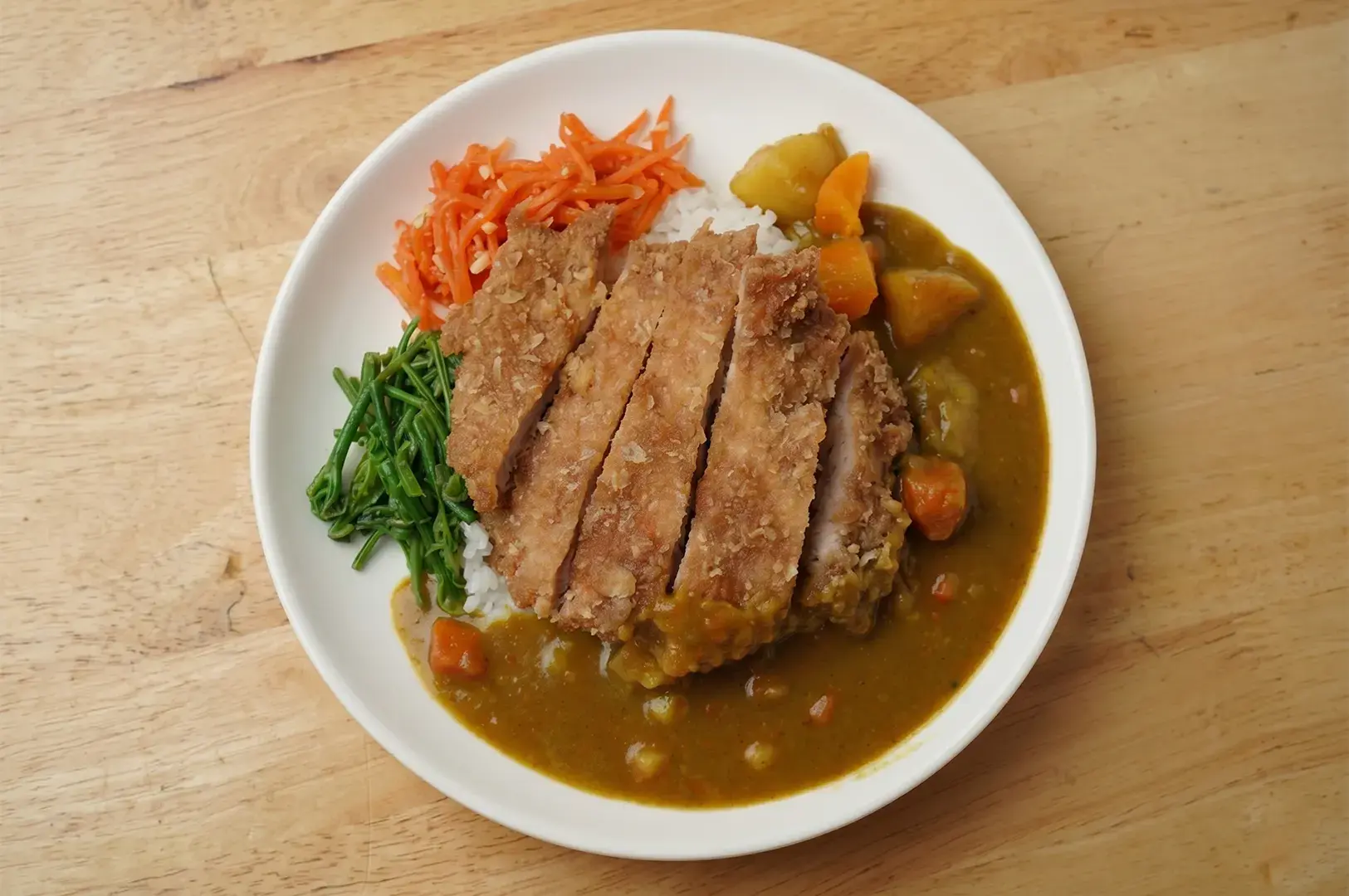Japanese chicken katsu curry rice plate with sliced crispy cutlet, vegetable side dishes, and potatoes on a wooden table.