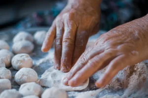 Close-up of elderly hands flattening dough into small balls on a floured surface.