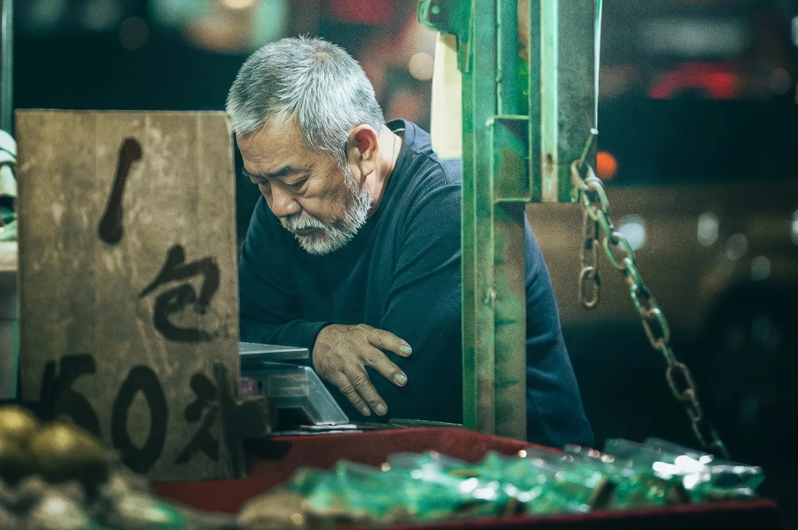 Candid street photography of an elderly street vendor with a gray beard looking down thoughtfully at his stall in a night market, featuring cinematic lighting and shallow depth of field.