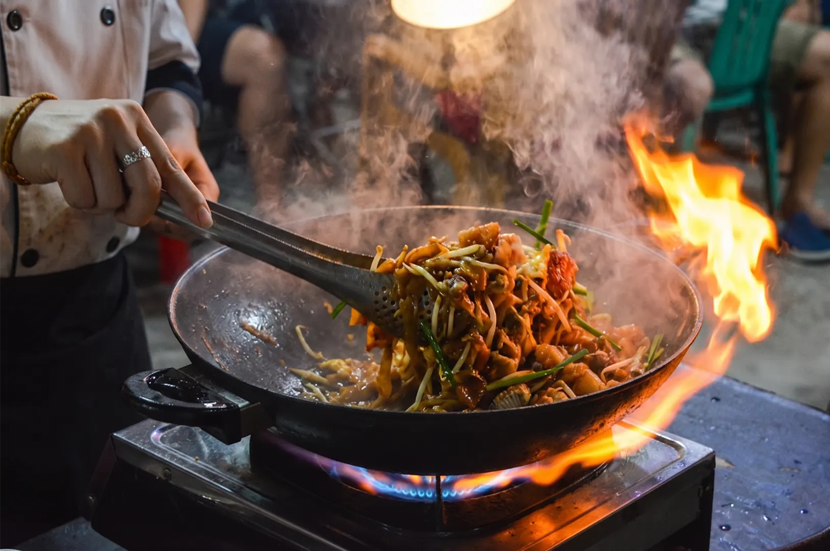 Professional chef stir-frying in a wok with a large burst of fire, demonstrating the "wok hei" technique in an outdoor kitchen at dusk.