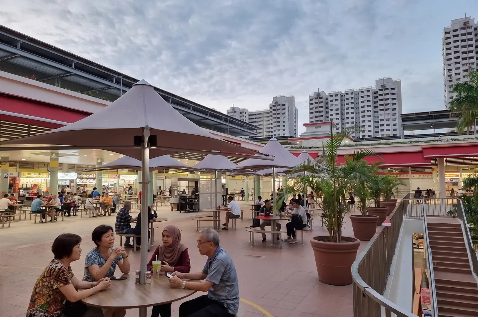 People dining at an open-air hawker center with umbrella-shaded tables and high-rise apartments in the background.