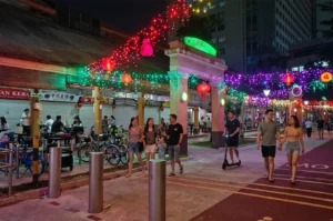 Pedestrians walking through a vibrant outdoor night market decorated with red Chinese lanterns and colorful festive fairy lights.