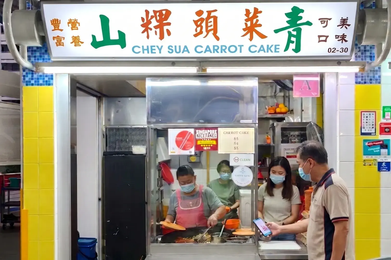 Chey Sua Carrot Cake hawker stall in Singapore with staff cooking on a flat-top grill and a customer paying using a smartphone.