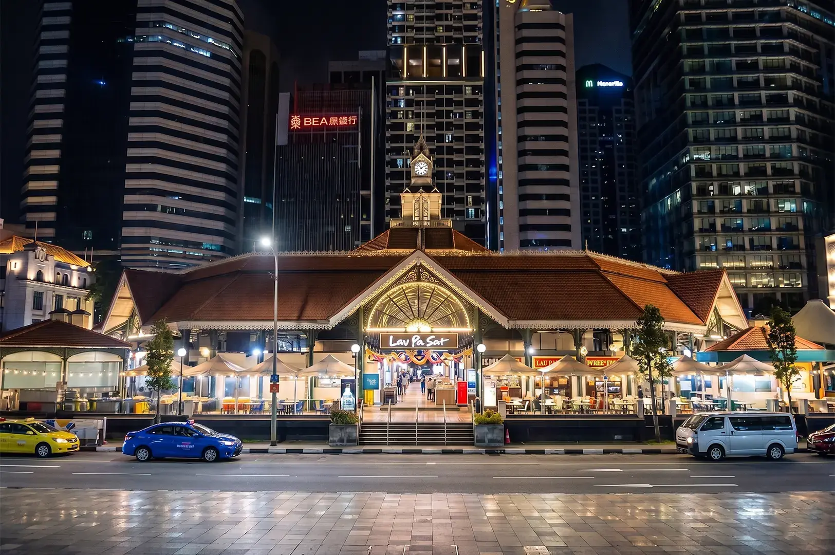Exterior view of the historic Lau Pa Sat hawker center at night, illuminated against the modern skyscrapers of Singapore's central business district.