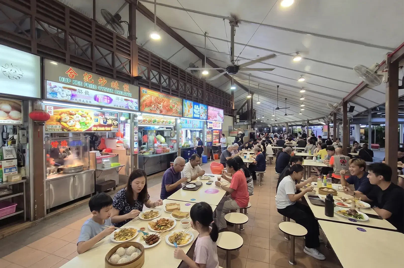 Families and groups dining at an indoor Singaporean hawker center with food stalls like Hup Kee Fried Oyster Omelette.