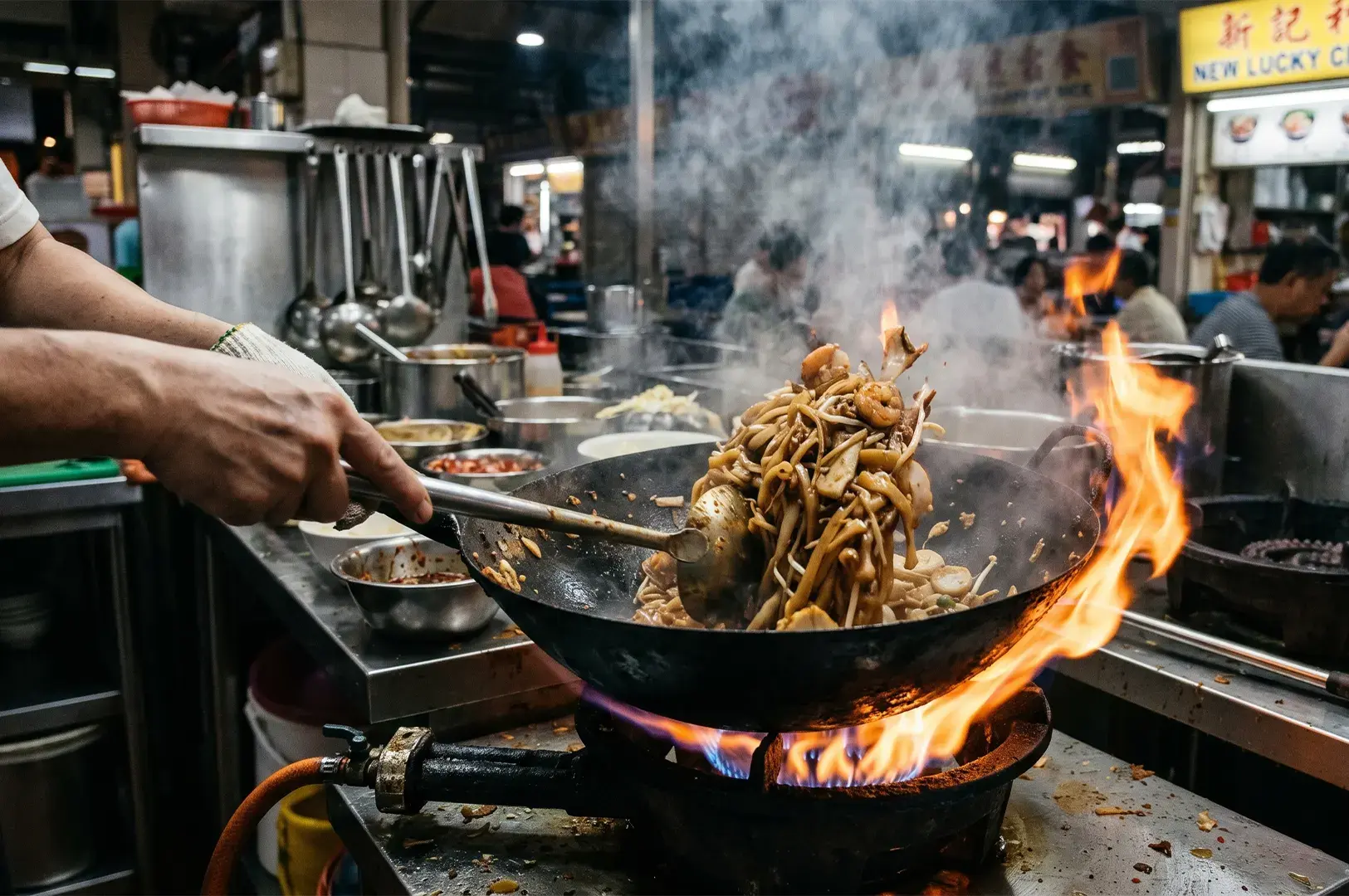 A chef stir-frying noodles and shrimp in a large wok with flames over a high-heat gas burner at a Singaporean hawker stall.