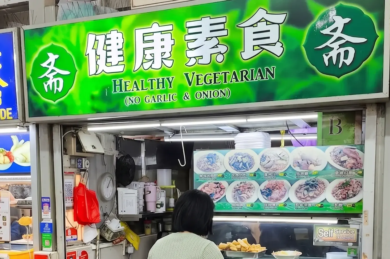 Healthy Vegetarian hawker stall storefront with a green sign indicating "No Garlic & Onion" and a menu display of Chinese vegetarian dishes.