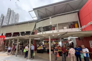 An open-air market and hawker center in Toa Payoh, Singapore, with people walking and shopping under a covered walkway, set against a backdrop of modern high-rise HDB flats.