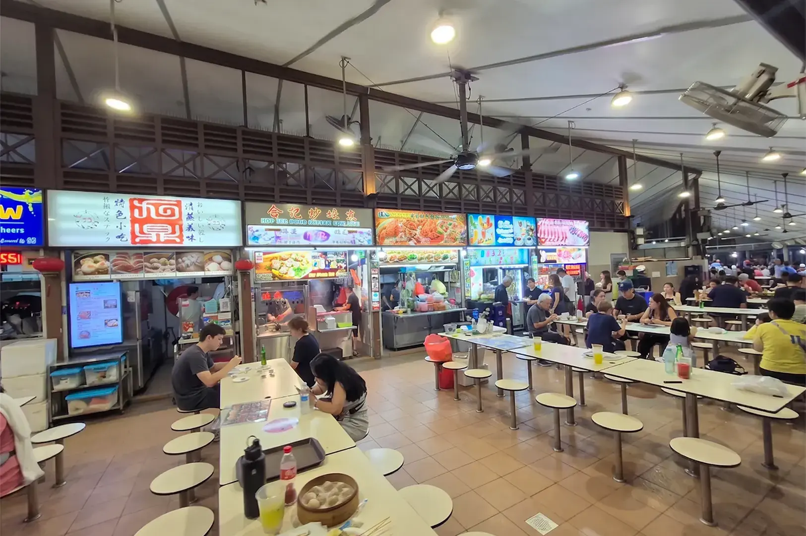 Patrons dining at an outdoor hawker center in Singapore at night, featuring various food stalls and communal seating.
