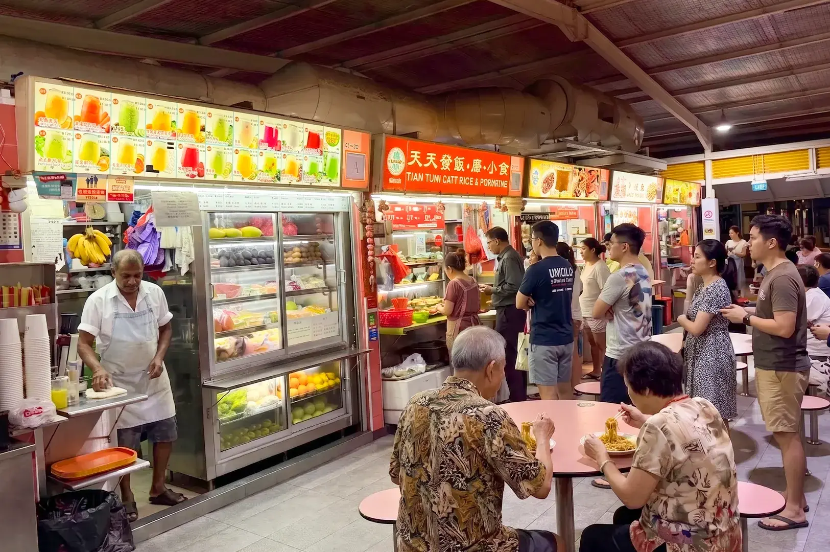 A busy outdoor hawker center food stall in Singapore with people lining up to order and customers eating noodles at a round table.
