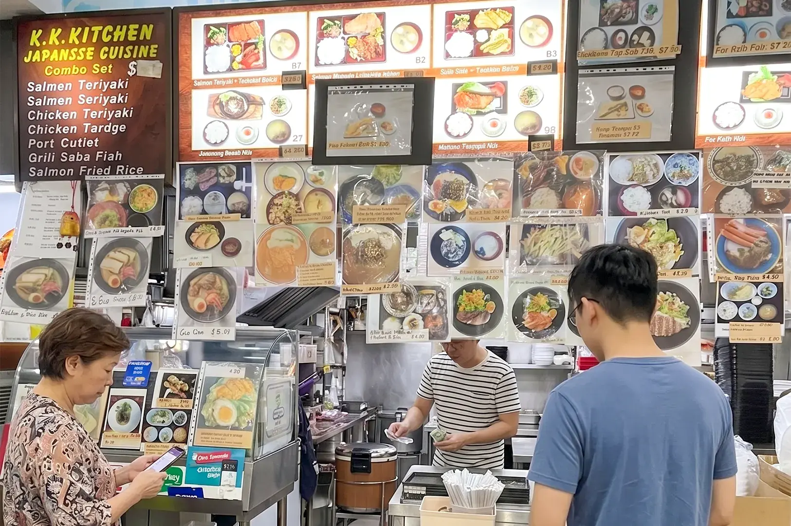 K.K. Kitchen Japanese Cuisine hawker stall storefront with a detailed menu display and customers ordering.