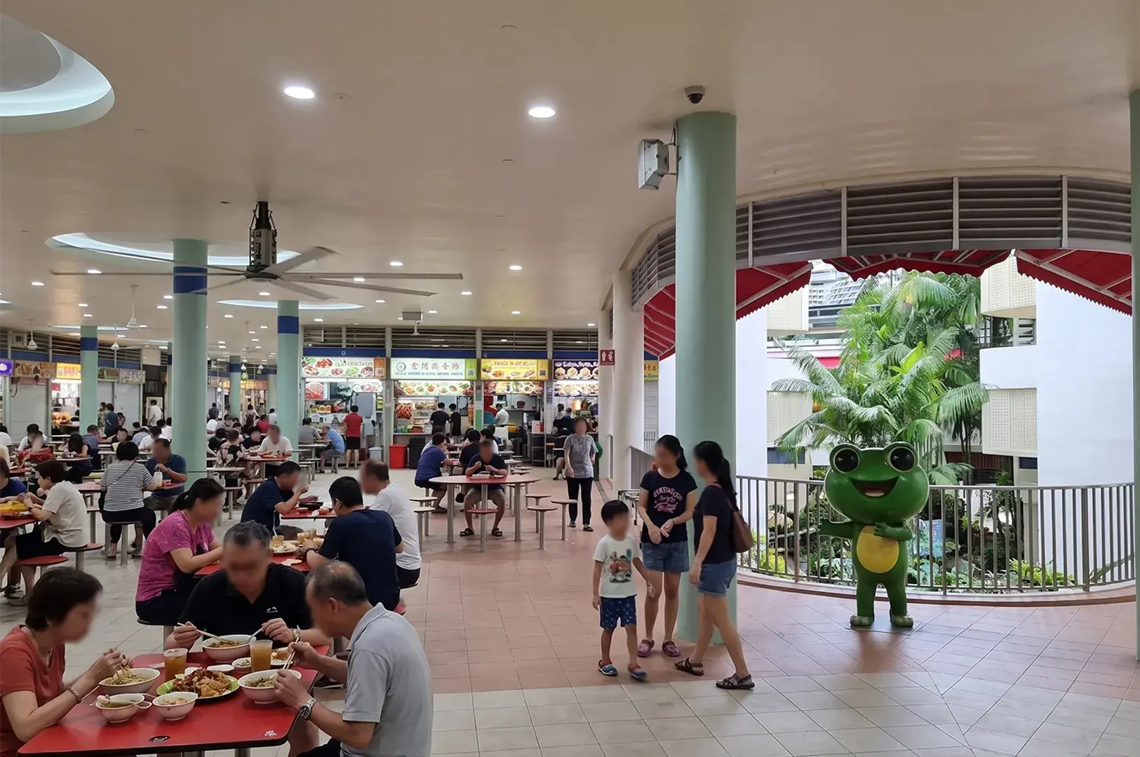 Families dining at a busy indoor hawker center with various food stalls and a green frog mascot statue.