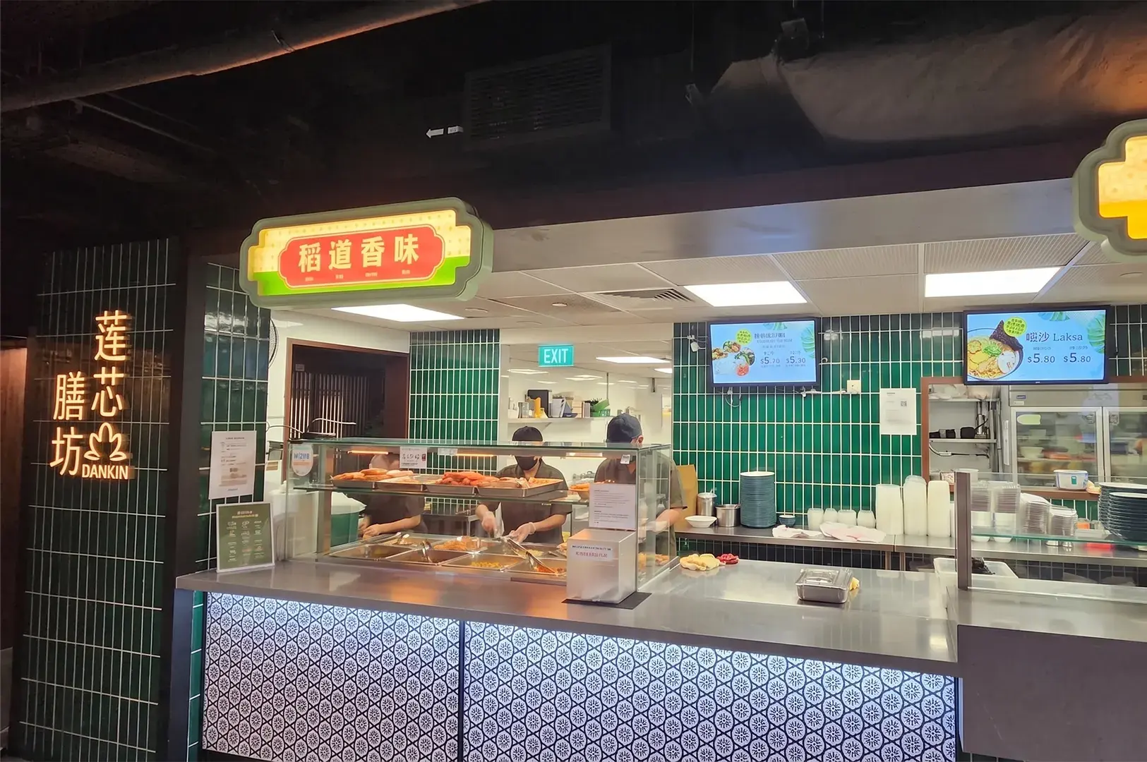 Dankin vegetarian food stall counter featuring a green tiled wall, digital menu screens for laksa, and staff preparing food.