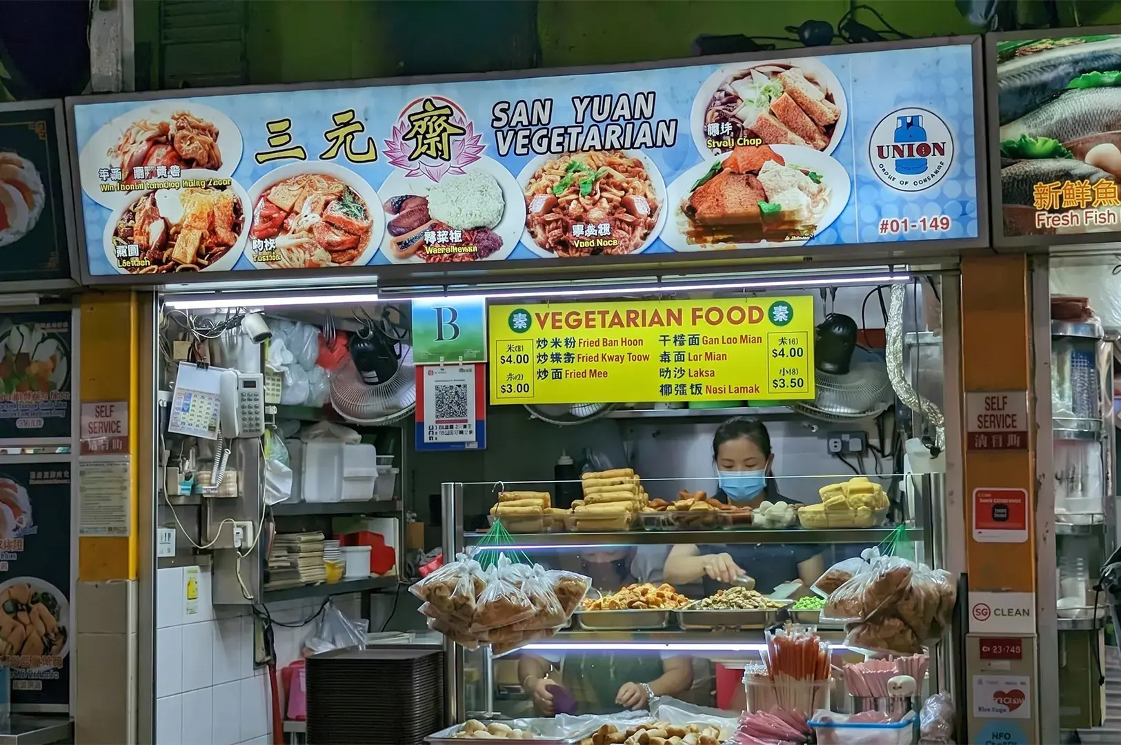 San Yuan Vegetarian hawker stall storefront with a blue menu board displaying Chinese vegetarian dishes like laksa, fried mee, and nasi lemak.