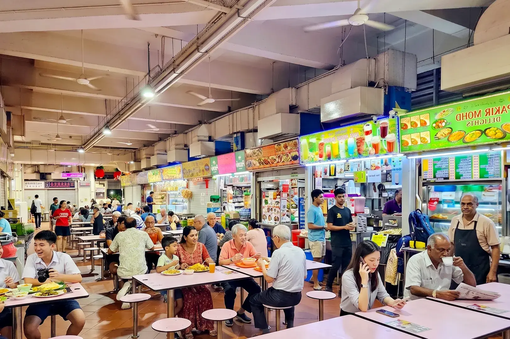 Crowded indoor hawker center food court in Singapore with people dining at tables near various local food stalls.