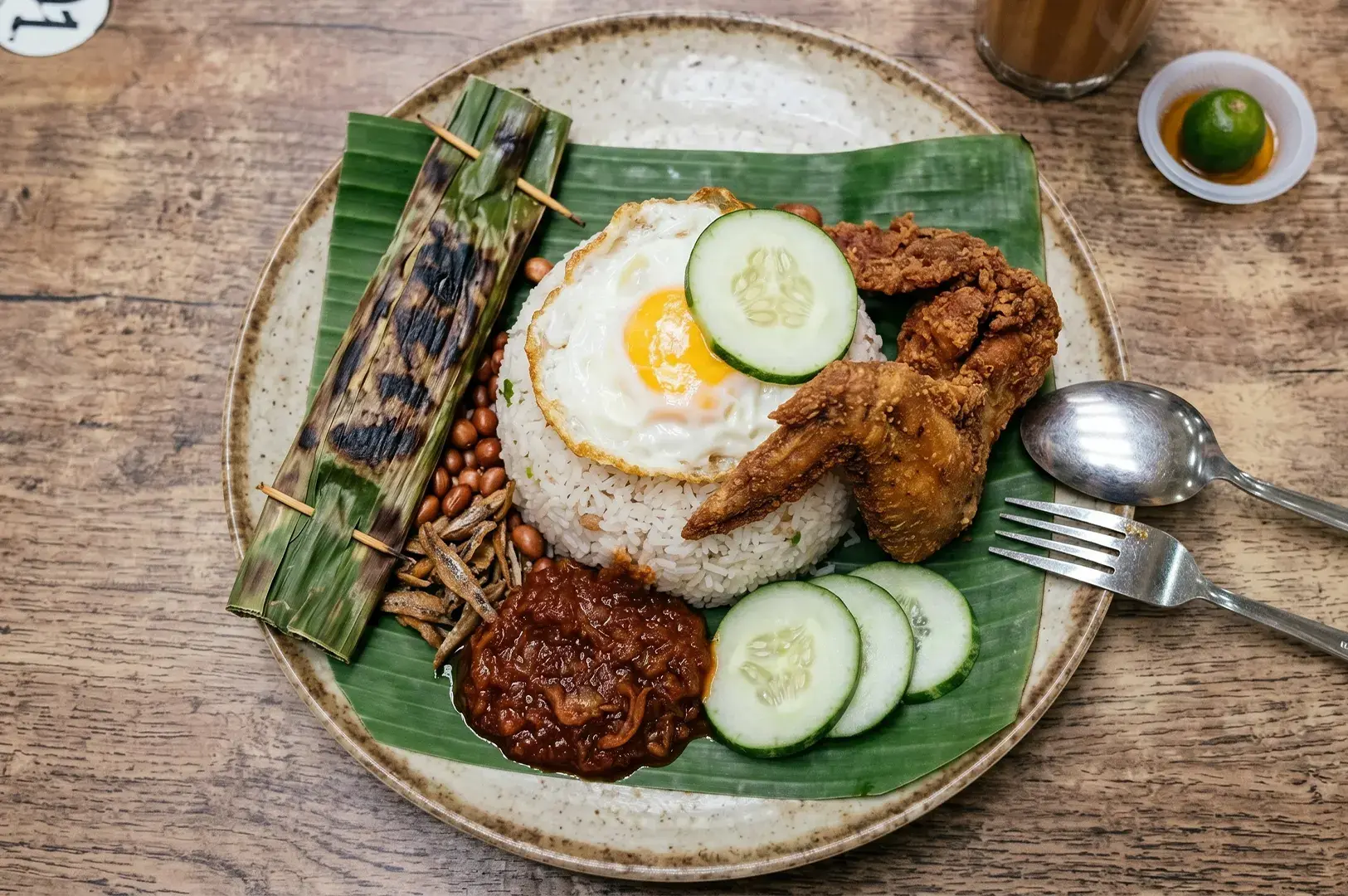 Nasi Lemak featuring fried chicken, a fried egg, sambal, anchovies, peanuts, cucumber slices, and grilled otak-otak served on a banana leaf.