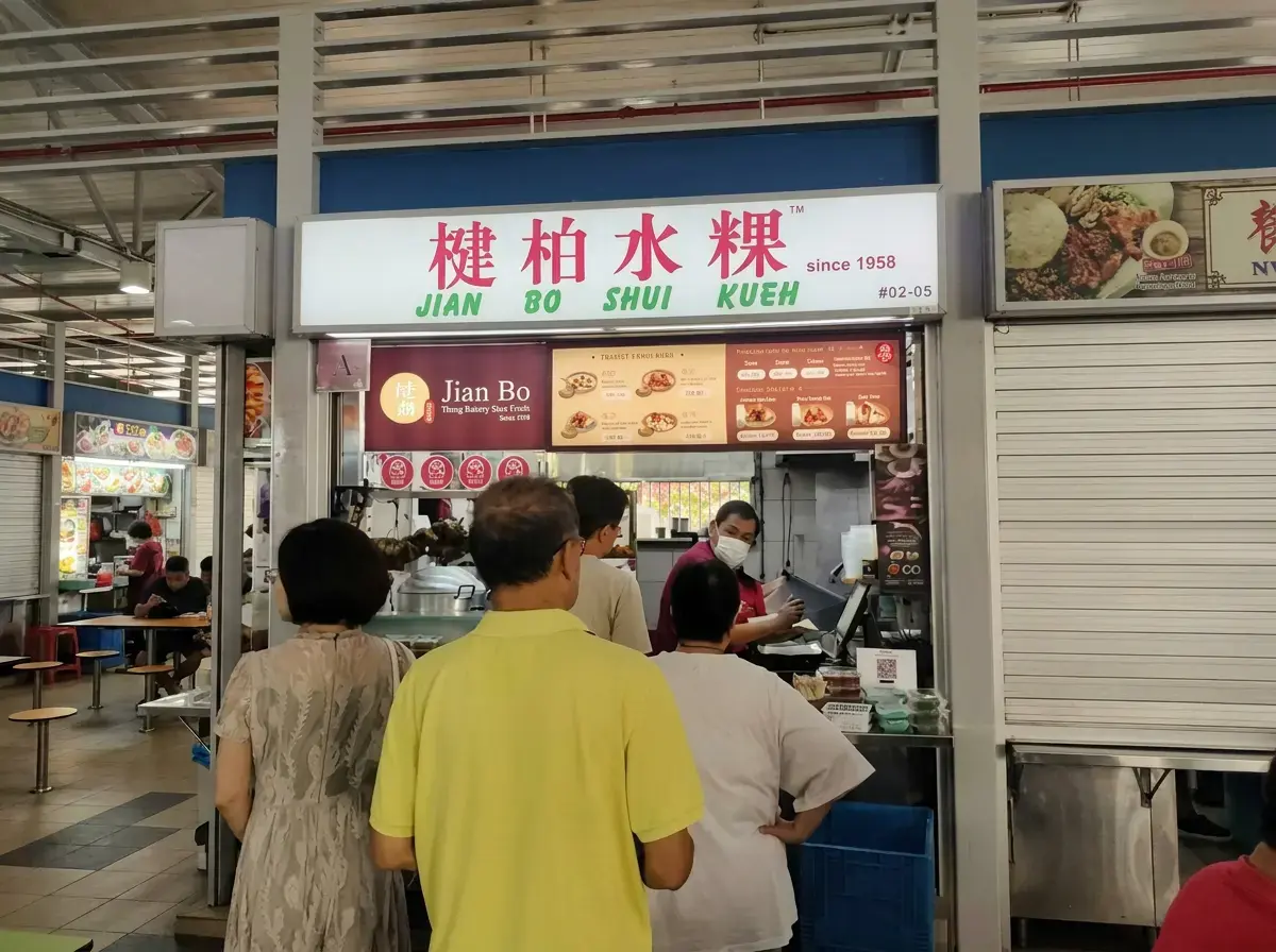 A line of customers waiting to order at the storefront counter of a "Jian Bo Shui Kueh" food stall in a hawker center.