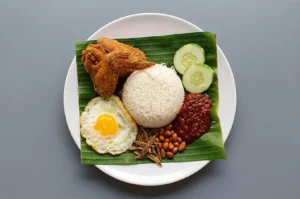 Nasi Lemak served on a banana leaf featuring coconut rice, fried chicken, a fried egg, sambal, peanuts, anchovies, and cucumber slices on a white plate.