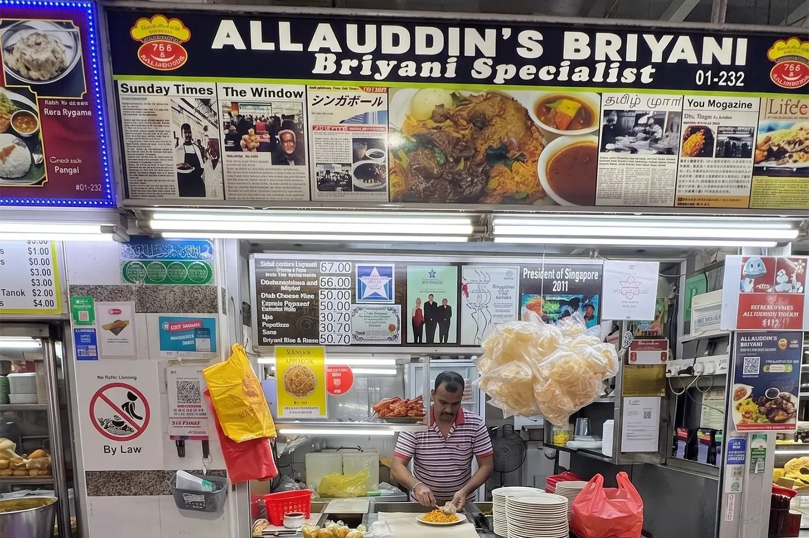 Allauddin's Briyani hawker stall storefront featuring a menu board covered in newspaper clippings and a vendor plating food.