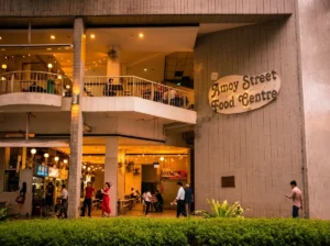 An exterior view of the multi-story Amoy Street Food Centre under warm evening lights, with people walking by and dining on the open balconies.