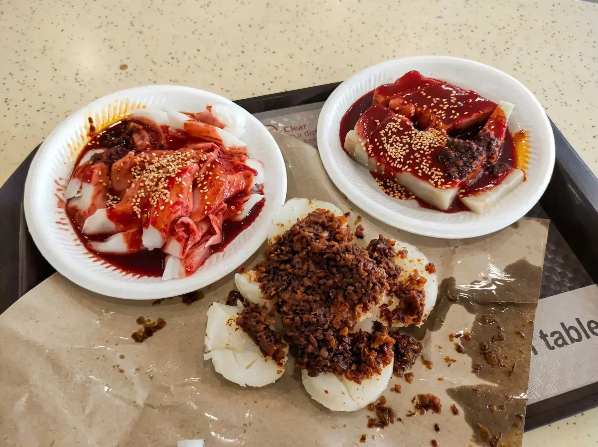 A food tray holding three local dishes: steamed rice cakes topped with preserved radish on brown paper, steamed rice noodle rolls in red sauce, and sliced yam cake in red sauce, both garnished with sesame seeds.