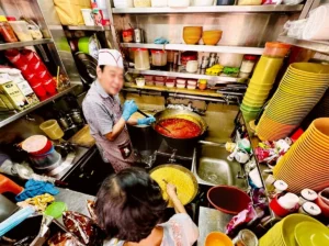 An overhead view inside a busy, cramped commercial kitchen where staff are preparing food in large pots surrounded by shelves of bowls and supplies.