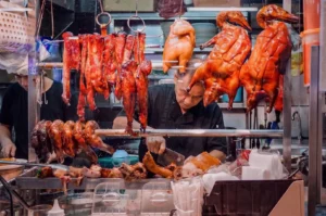 Hawker chef chopping roasted meat behind a display window hanging with char siu and roasted ducks.
