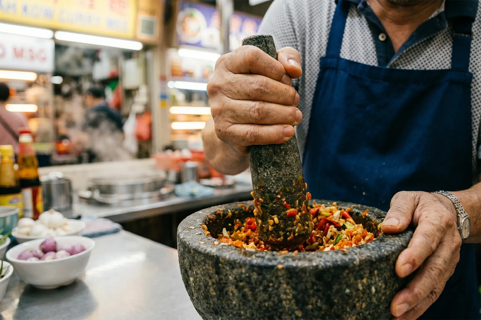 Hawker chef using a stone mortar and pestle to grind fresh chili paste sambal at a food stall.