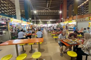 People dining at tables inside a busy Singapore hawker center with food stalls.