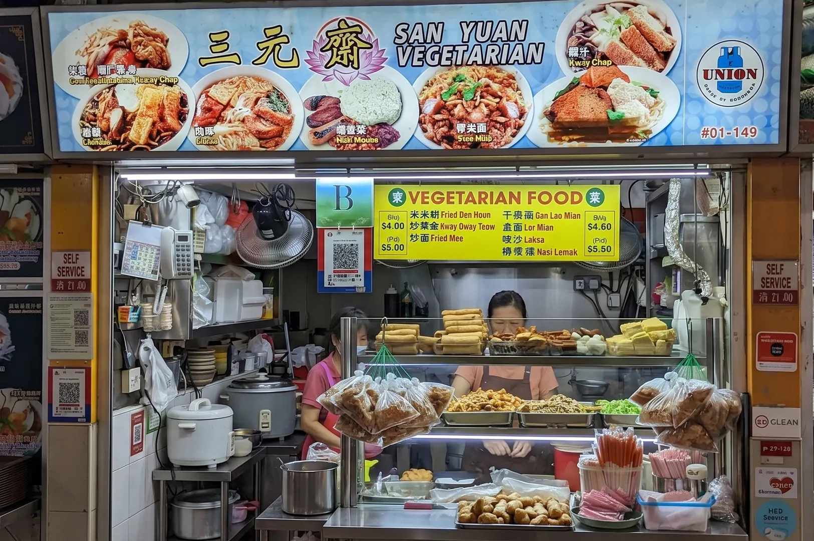 San Yuan Vegetarian food stall at a Singapore hawker center with a colorful menu banner and display of dishes.