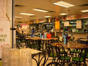 Indoor view of a traditional food court or cooked food centre featuring large round wooden dining tables paired with green and black metal chairs, set against a backdrop of Chinese food stalls under warm fluorescent lighting.