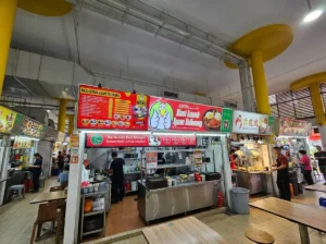 A straight-on view of the "Nasi Lemak Ayam Taliwang" food stall in a hawker center, featuring a bright red menu board and a banner highlighting its Michelin rating.