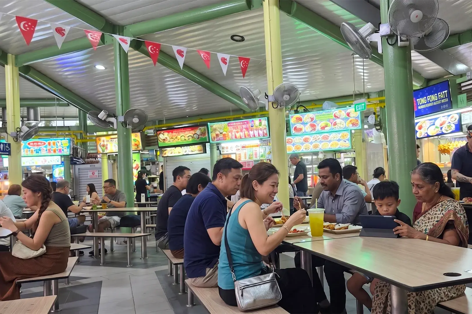 People dining together inside a busy Singapore hawker center decorated with small flags.