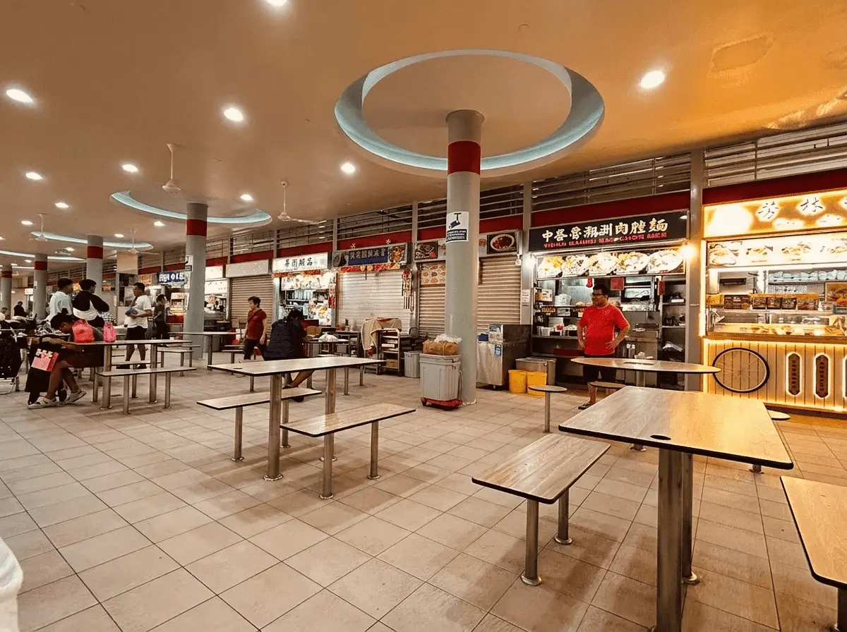Interior view of a brightly lit, spacious Singapore hawker centre with rows of empty wooden tables and benches, large structural pillars, and various food stalls in the background.