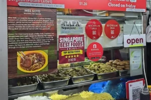A close-up of a hawker stall food counter displaying ingredients, Michelin Bib Gourmand stickers, and an information board describing traditional Mee Soto.