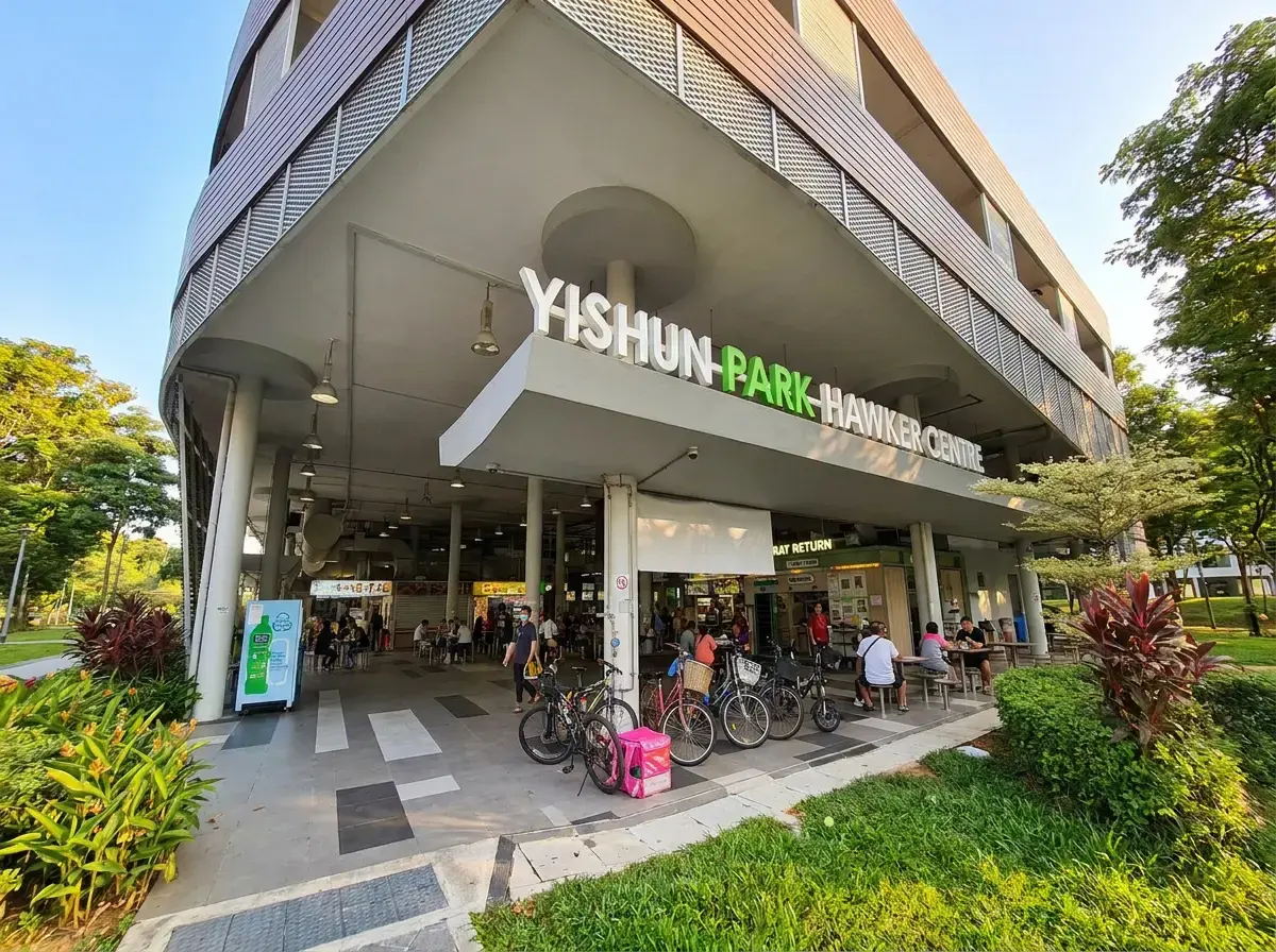 A wide shot of the modern exterior entrance to "Yishun Park Hawker Centre," featuring large white signage, parked bicycles, and open-air seating areas.