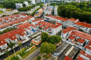 An aerial view of a low-rise residential complex with distinct orange-red roofs and white walls, nestled alongside lush green trees and urban streets.