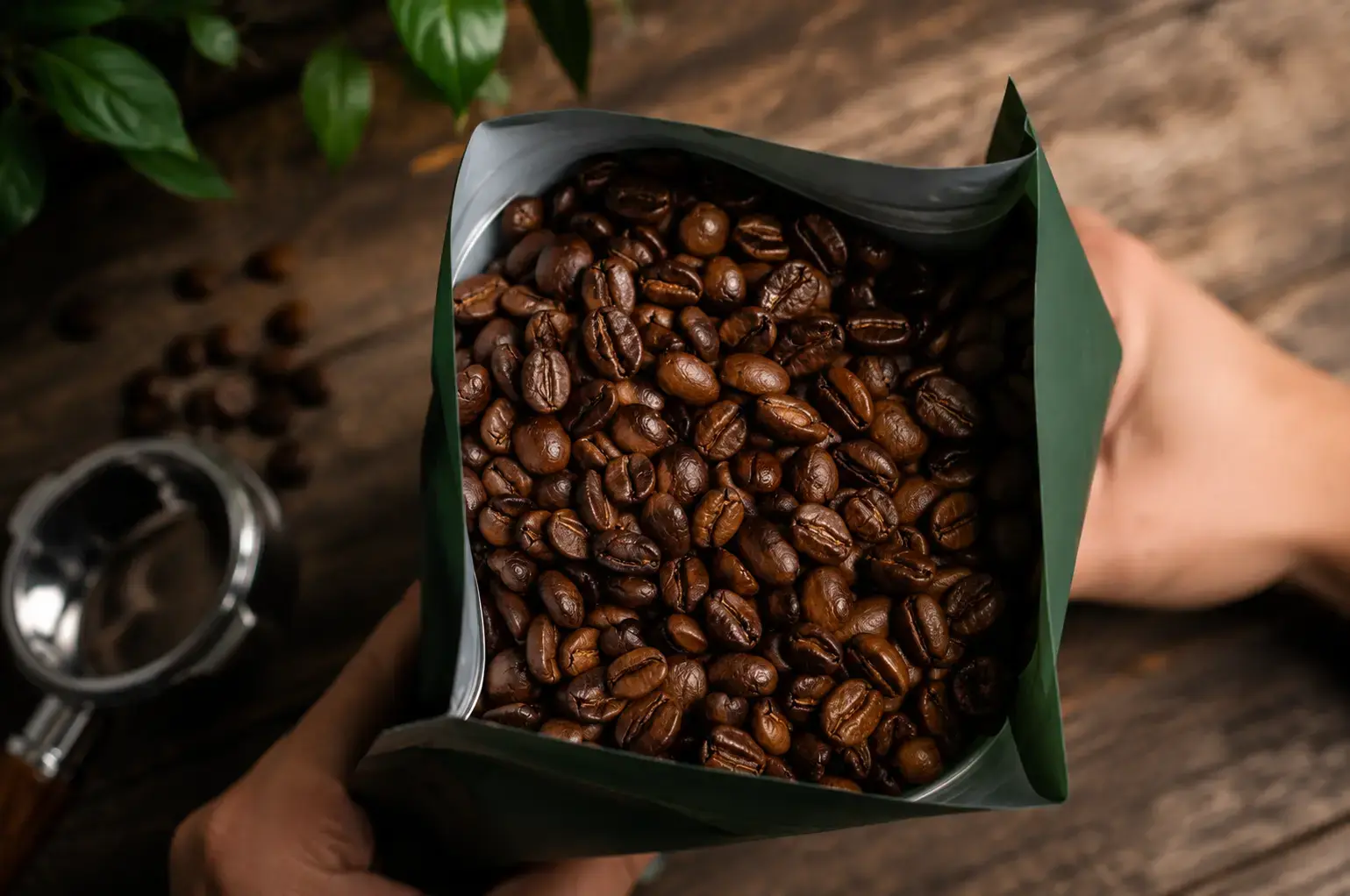 Top-down view of hands holding open a dark green bag filled with roasted coffee beans on a rustic wooden table.