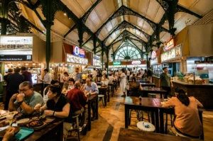 A bustling indoor food hall with a high, vaulted ceiling and intricate green metal arches, filled with people sitting at wooden tables and eating from various food stalls.