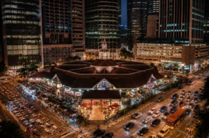 An aerial night view of the octagonal Lau Pa Sat hawker center in Singapore, nestled among glowing skyscrapers and surrounded by busy streets with outdoor dining.